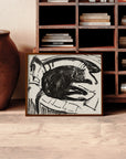 A framed "Lying Cat" print, featuring an abstract black and white design, is propped against a wooden bookshelf. The shelf is filled with various books stacked both horizontally and vertically, embodying an artistic vision. A large clay pot sits next to the artwork on a light wood floor.
fb-feed