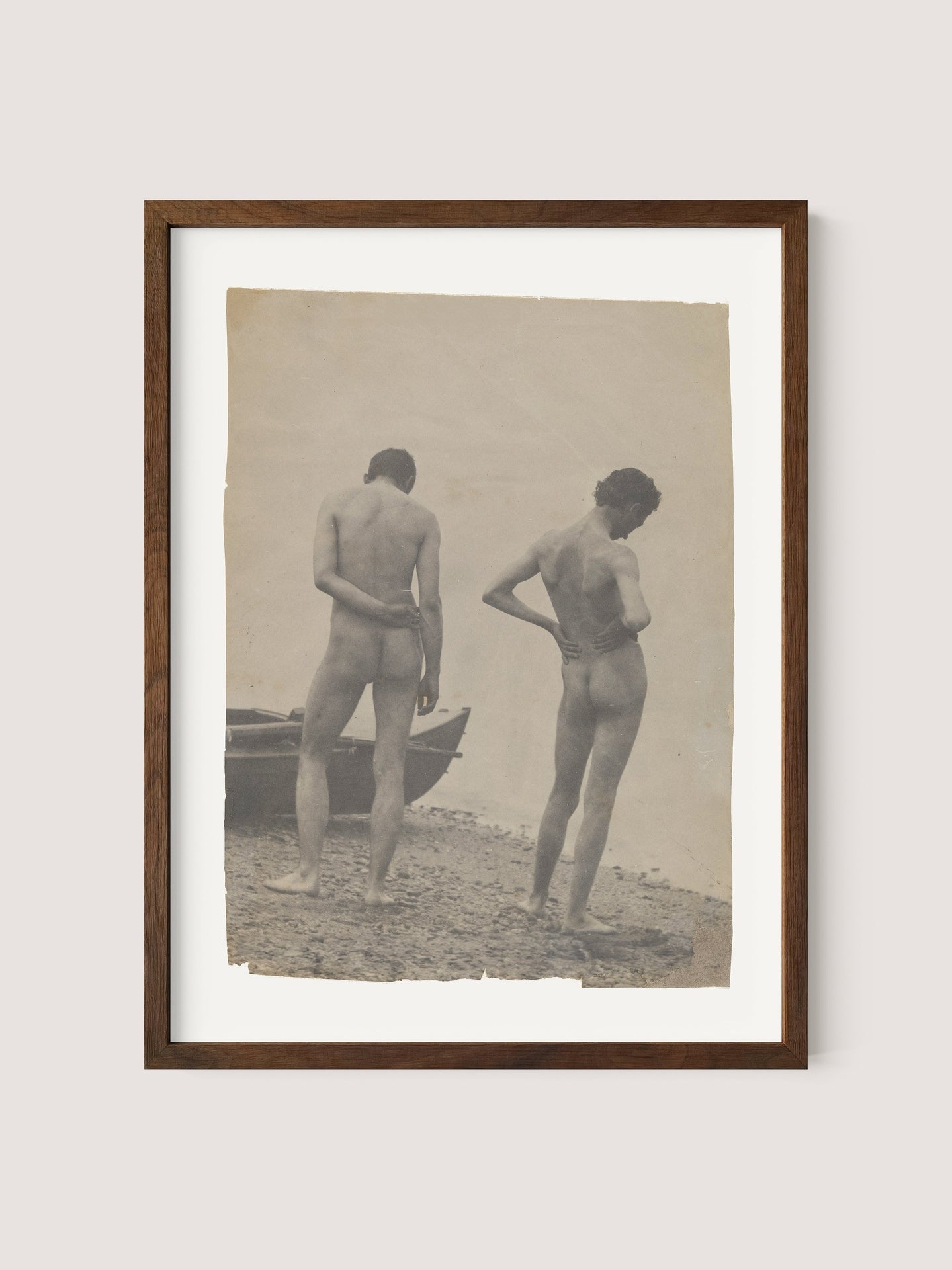 The framed sepia-toned photograph titled "Beach Walk," evocative of Thomas Eakins' artistry, features two nude figures standing on a rocky shoreline. Both face away from the camera; one holds their left arm behind their back while the other places their right hand on their hip. A wooden boat is partially visible on the left, encapsulating a moment of creative photography, all set in a beautiful walnut oak frame.