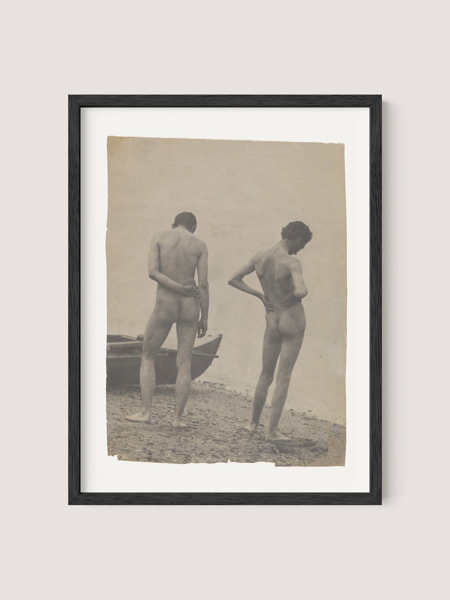 A framed vintage photograph titled "Beach Walk," reminiscent of Thomas Eakins' work, depicts two nude individuals standing on a beach. They face away from the camera, gazing toward the water with a small boat to their left. The image's sepia tone enhances its aged appearance, reflecting a timeless creative process in photography. The frame itself is finished in Black Oak, adding an elegant touch to this evocative piece of art.