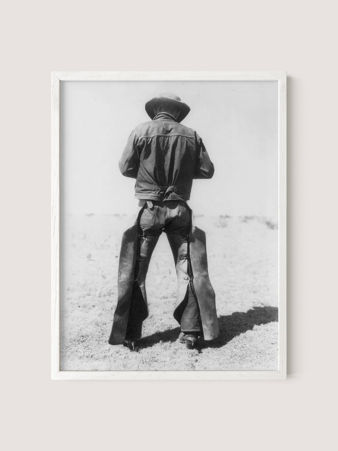 The "Working Cowboy" is a black and white photograph framed in white oak, showcasing an American cowboy from behind. He is wearing a cowboy hat, a long-sleeved shirt, and chaps, standing in a barren, sandy landscape. This nostalgic image captures the figure gazing into the distance.