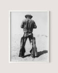 The "Working Cowboy" is a black and white photograph framed in white oak, showcasing an American cowboy from behind. He is wearing a cowboy hat, a long-sleeved shirt, and chaps, standing in a barren, sandy landscape. This nostalgic image captures the figure gazing into the distance.