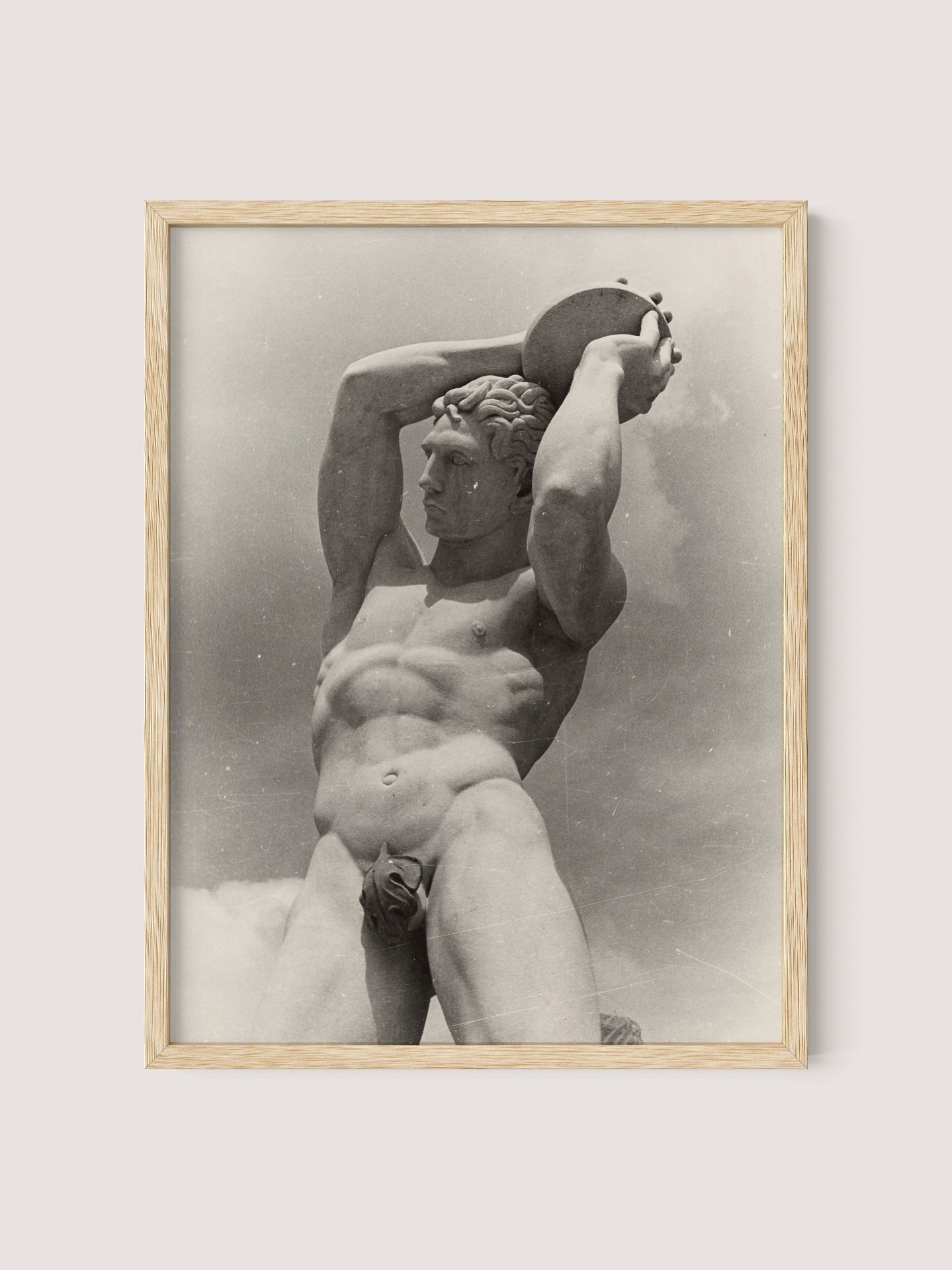 A framed photograph titled "The Discus Thrower" by Paul Wolff features a nude male statue holding a disc above his head with both hands. The sepia-toned image, set against a cloudy sky, exudes a classic and timeless feel, complemented by its natural oak frame.