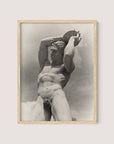 A framed photograph titled "The Discus Thrower" by Paul Wolff features a nude male statue holding a disc above his head with both hands. The sepia-toned image, set against a cloudy sky, exudes a classic and timeless feel, complemented by its natural oak frame.