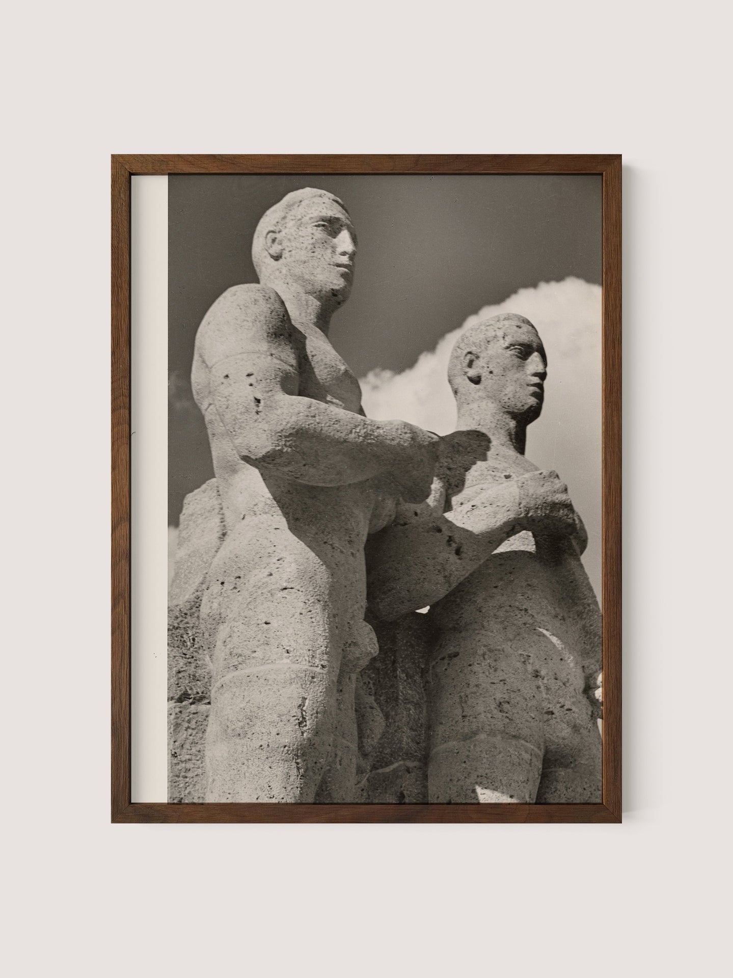 A framed black and white photograph titled &quot;Olympic Men, Berlin&quot; by Paul Wolff shows a sculpted stone relief of two muscular, stoic figures standing side by side, with one slightly in front of the other. The sky with clouds forms the background, reminiscent of Berlin Olympic statues from the 1936 Olympic Games. The frame is simple and crafted from walnut oak.