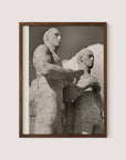 A framed black and white photograph titled "Olympic Men, Berlin" by Paul Wolff shows a sculpted stone relief of two muscular, stoic figures standing side by side, with one slightly in front of the other. The sky with clouds forms the background, reminiscent of Berlin Olympic statues from the 1936 Olympic Games. The frame is simple and crafted from walnut oak.