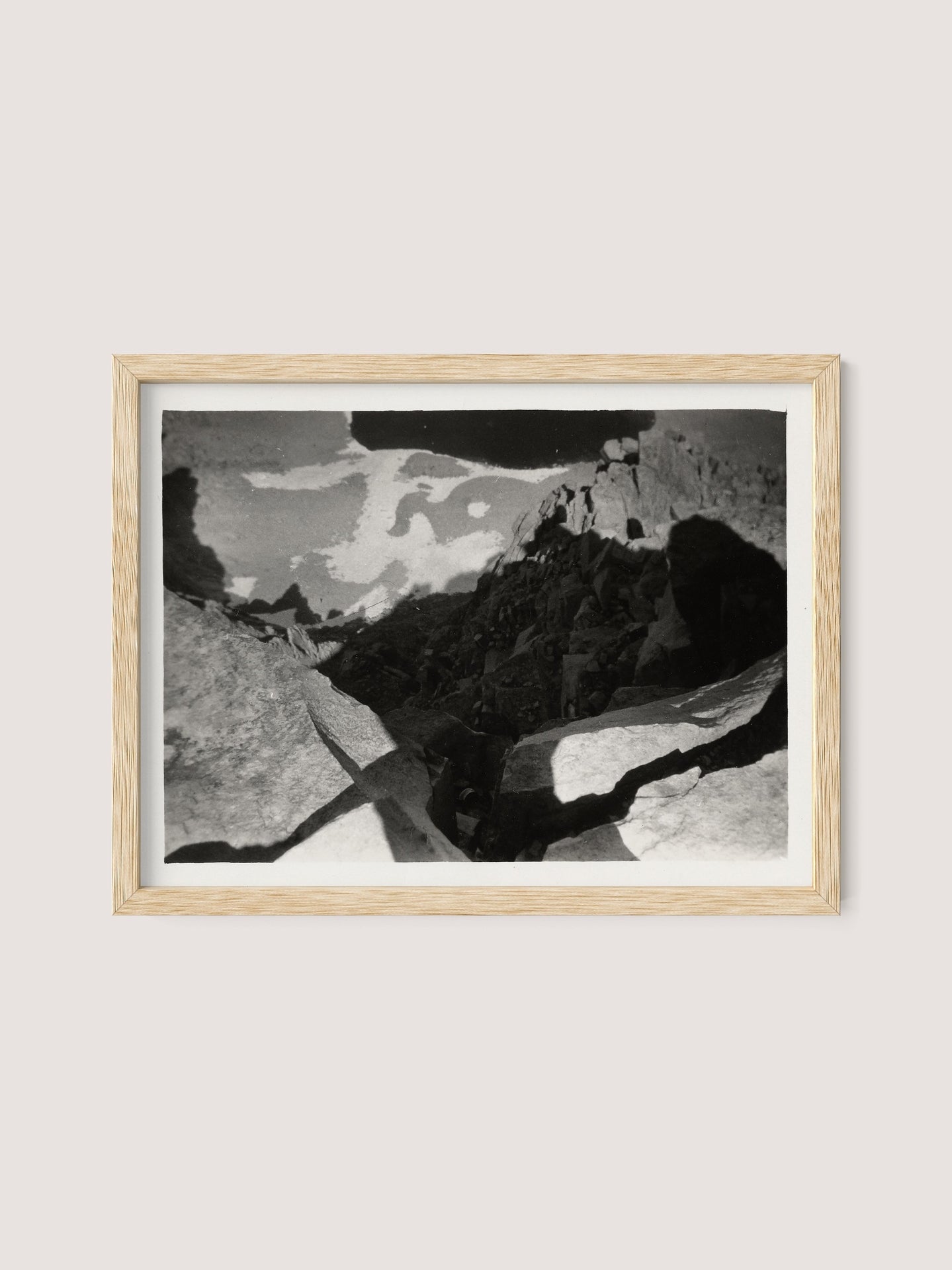 A framed black-and-white photograph titled "Looking Down From Mt. Whitney" by Louis Fleckenstein depicts the rocky terrain of Mt. Whitney with scattered shadows. The image has a surreal, abstract quality, emphasizing the dramatic interplay of light and darkness in the scene. The frame is made of natural oak wood.