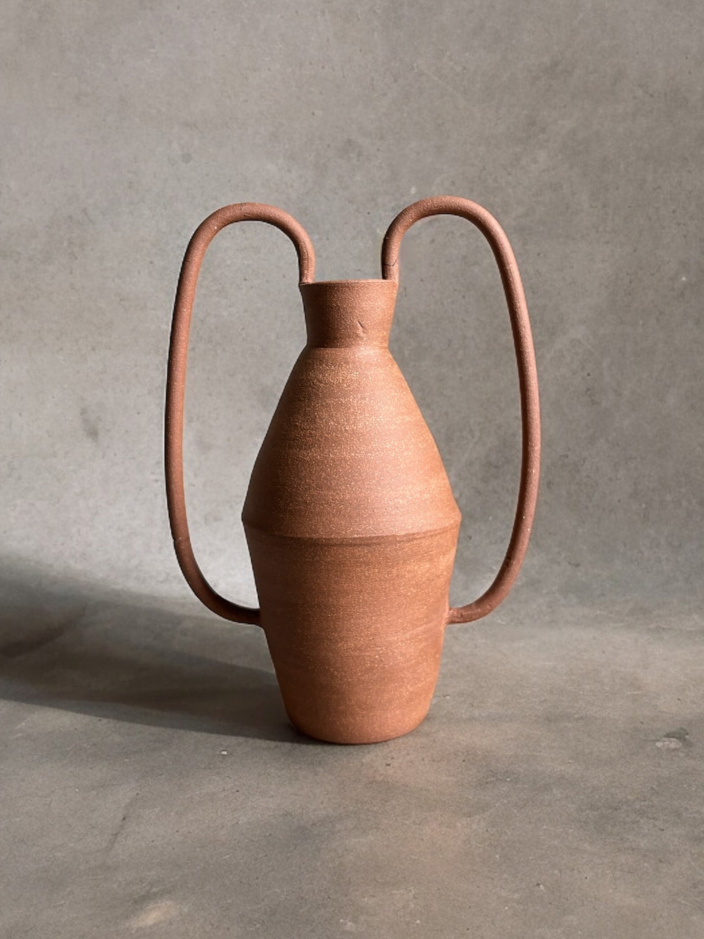 The Amphora Vase is a handcrafted ceramic piece with a terracotta finish, featuring a tall, narrow body and two large, U-shaped handles extending from the neck to the base. It is set against a plain, neutral background.