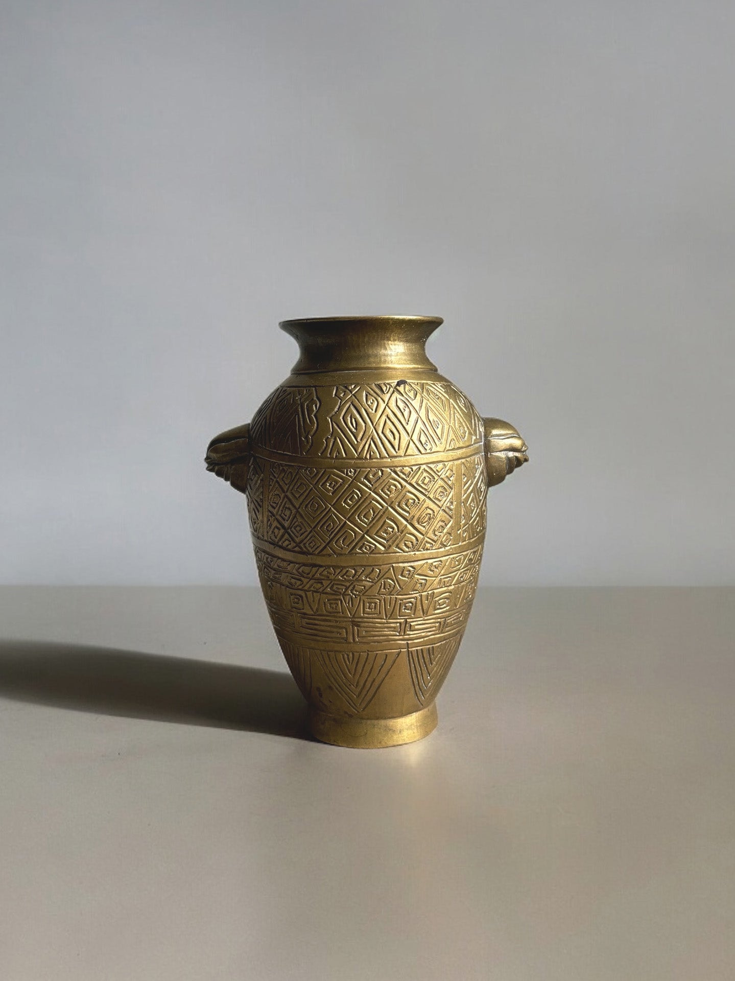 An Egyptian Urn crafted from golden brass, featuring intricate geometric patterns of triangles and diamond shapes, adorned with two small handles on either side, set against a plain gray background. The urn has a gleaming finish.