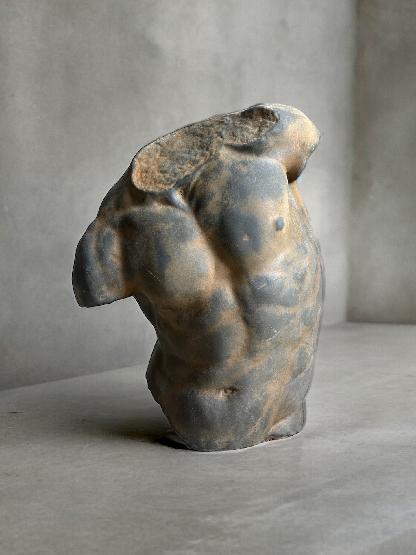 A captivating Plaster Torso sculpture of a male torso showcased on a smooth, light-colored surface. The artwork is headless and armless, highlighting its muscular definition and form. The surface of the sculpture boasts an aged, textured appearance with a blend of gray and brown shades.