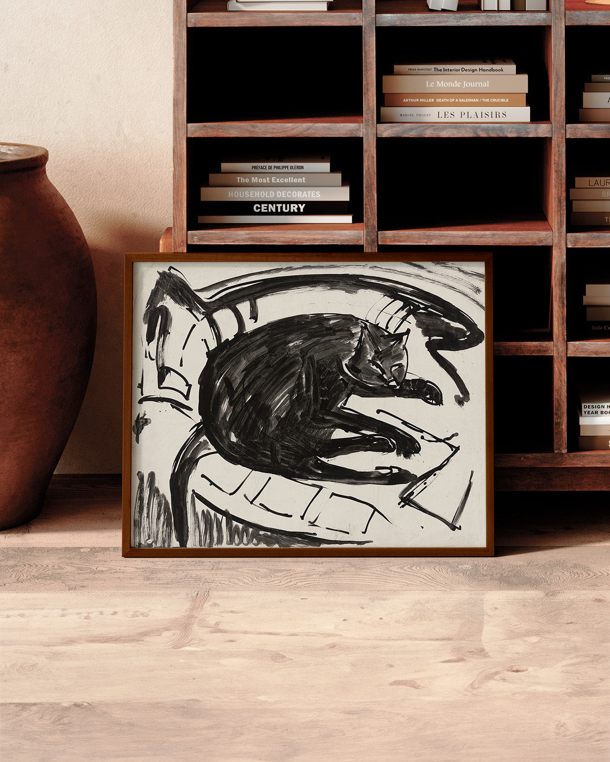 A framed "Lying Cat" print, featuring an abstract black and white design, is propped against a wooden bookshelf. The shelf is filled with various books stacked both horizontally and vertically, embodying an artistic vision. A large clay pot sits next to the artwork on a light wood floor.