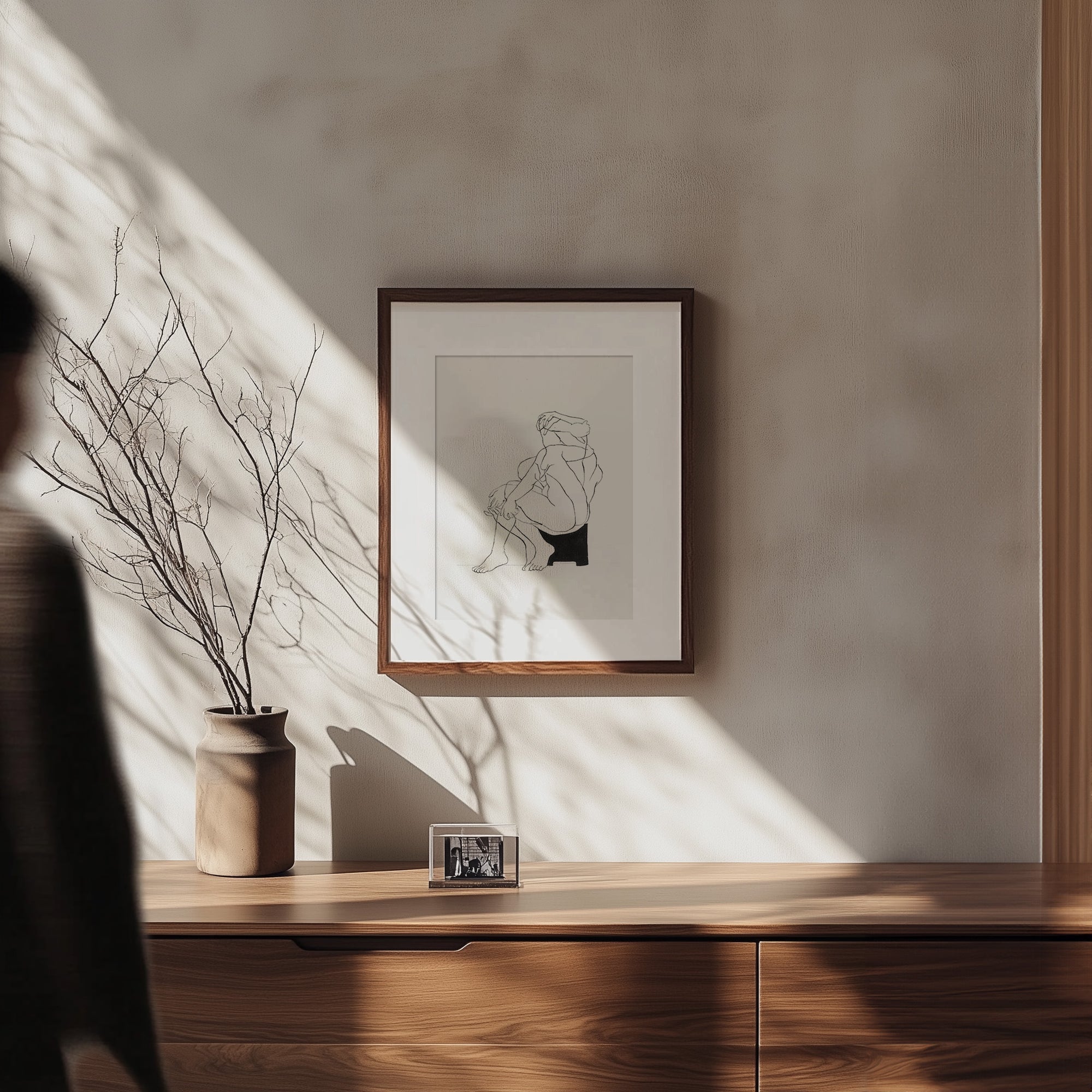 A person walks past the modern Samantha Michell II wooden sideboard, which is adorned with a potted plant of bare branches and a small framed photo. Sunlight streams in, casting shadows. Above the sideboard on the light-colored wall hangs an original minimalist ink drawing from an edition of 10.