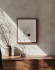 A person walks past the modern Samantha Michell II wooden sideboard, which is adorned with a potted plant of bare branches and a small framed photo. Sunlight streams in, casting shadows. Above the sideboard on the light-colored wall hangs an original minimalist ink drawing from an edition of 10.