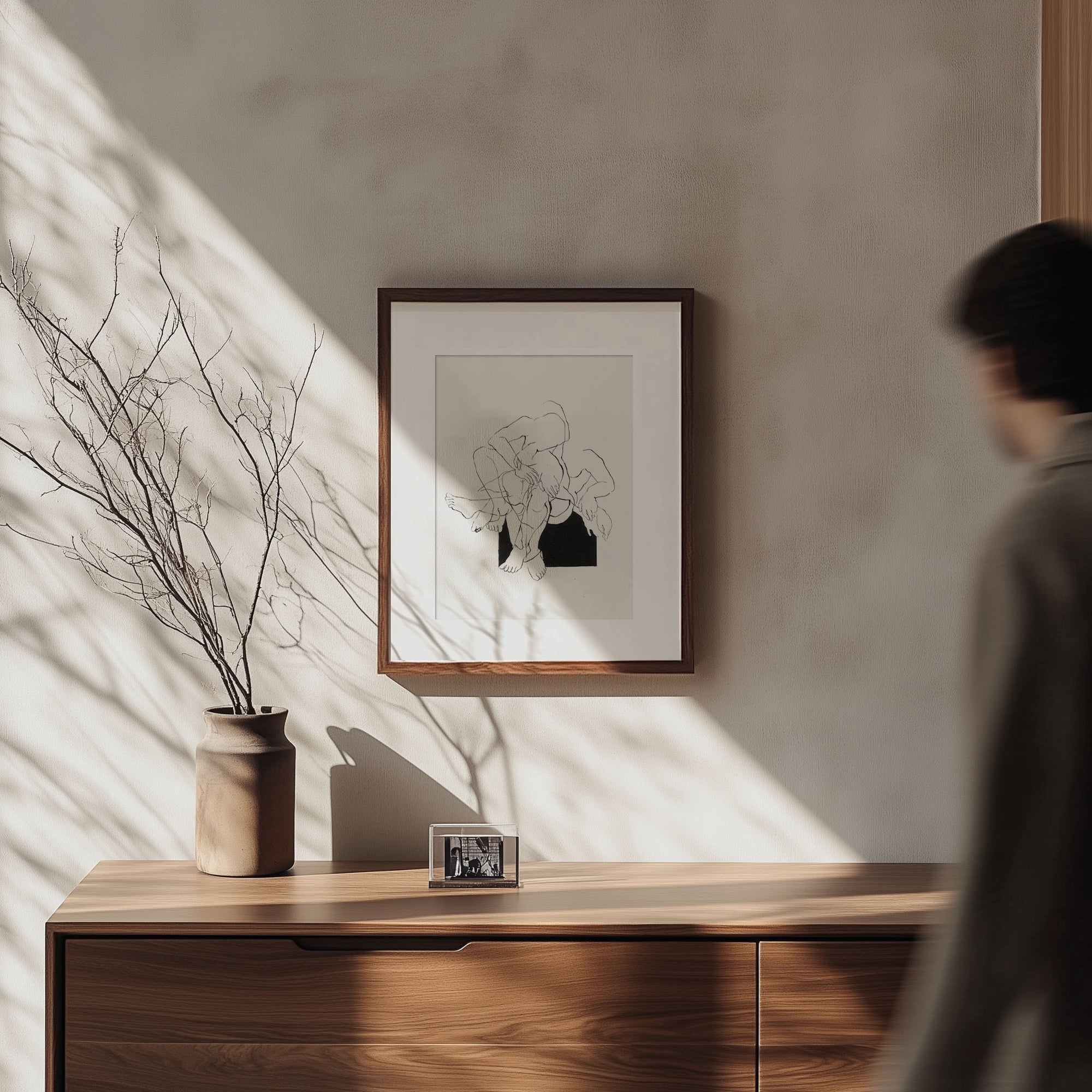 A minimalistic room with soft natural light illuminating a wooden sideboard featuring the Samantha Michell II. A framed abstract floral artwork, an original ink drawing, hangs above. A small vase with bare branches and a framed photograph sit on the sideboard as a person walks by, blurred in motion.