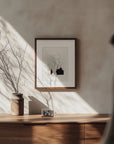 A minimalistic room with soft natural light illuminating a wooden sideboard featuring the Samantha Michell II. A framed abstract floral artwork, an original ink drawing, hangs above. A small vase with bare branches and a framed photograph sit on the sideboard as a person walks by, blurred in motion.