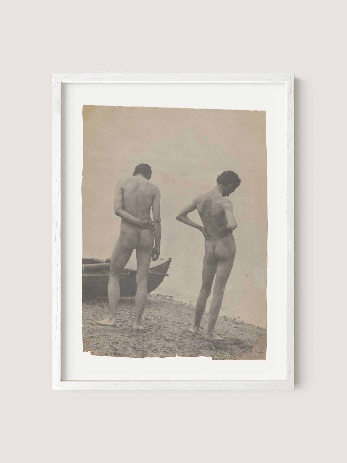 Beach Walk, a framed photograph with a sepia tone that echoes the artistic style of Thomas Eakins&#39; work, features two nude individuals standing on a rocky beach, their backs facing the camera. A boat is partially visible on the left side of the picture, enhancing its old, vintage allure. The frame comes in a refined White Oak color.