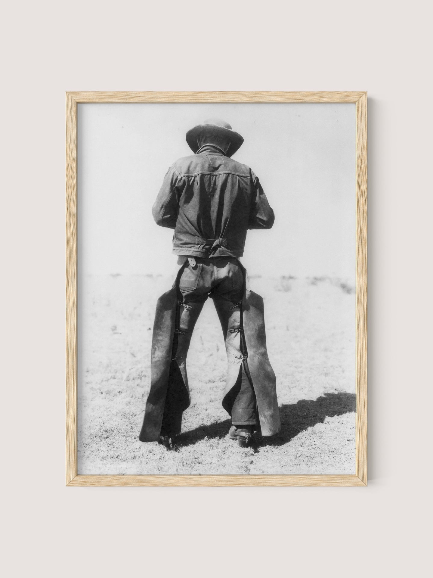 Natural Oak-framed photo titled "Working Cowboy," depicting an American Cowboy standing with his back to the camera. He is wearing a broad-brimmed hat, a denim shirt, and chaps. The nostalgic background appears to be a barren, dusty landscape.