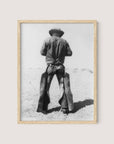 Natural Oak-framed photo titled "Working Cowboy," depicting an American Cowboy standing with his back to the camera. He is wearing a broad-brimmed hat, a denim shirt, and chaps. The nostalgic background appears to be a barren, dusty landscape.