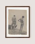 The framed sepia-toned photograph titled "Beach Walk," evocative of Thomas Eakins' artistry, features two nude figures standing on a rocky shoreline. Both face away from the camera; one holds their left arm behind their back while the other places their right hand on their hip. A wooden boat is partially visible on the left, encapsulating a moment of creative photography, all set in a beautiful walnut oak frame.