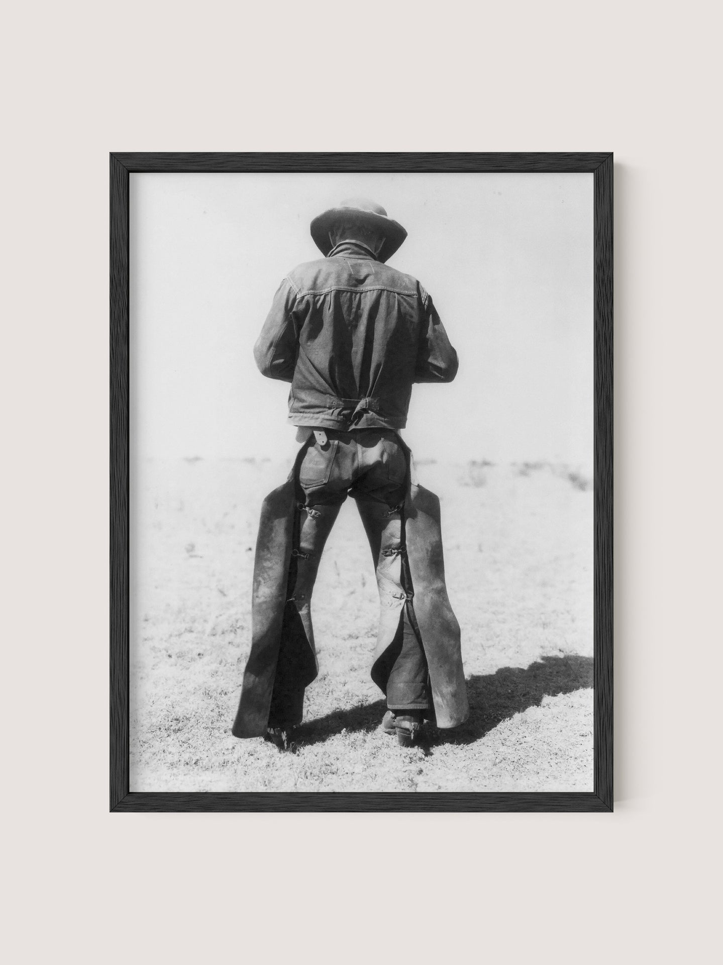 A nostalgic black and white photograph titled "Working Cowboy" features an American cowboy standing with his back to the camera. He is wearing a hat, a denim shirt, and chaps. The image is framed in a simple Black Oak frame, capturing an open, arid landscape in the background.