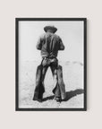 A nostalgic black and white photograph titled "Working Cowboy" features an American cowboy standing with his back to the camera. He is wearing a hat, a denim shirt, and chaps. The image is framed in a simple Black Oak frame, capturing an open, arid landscape in the background.