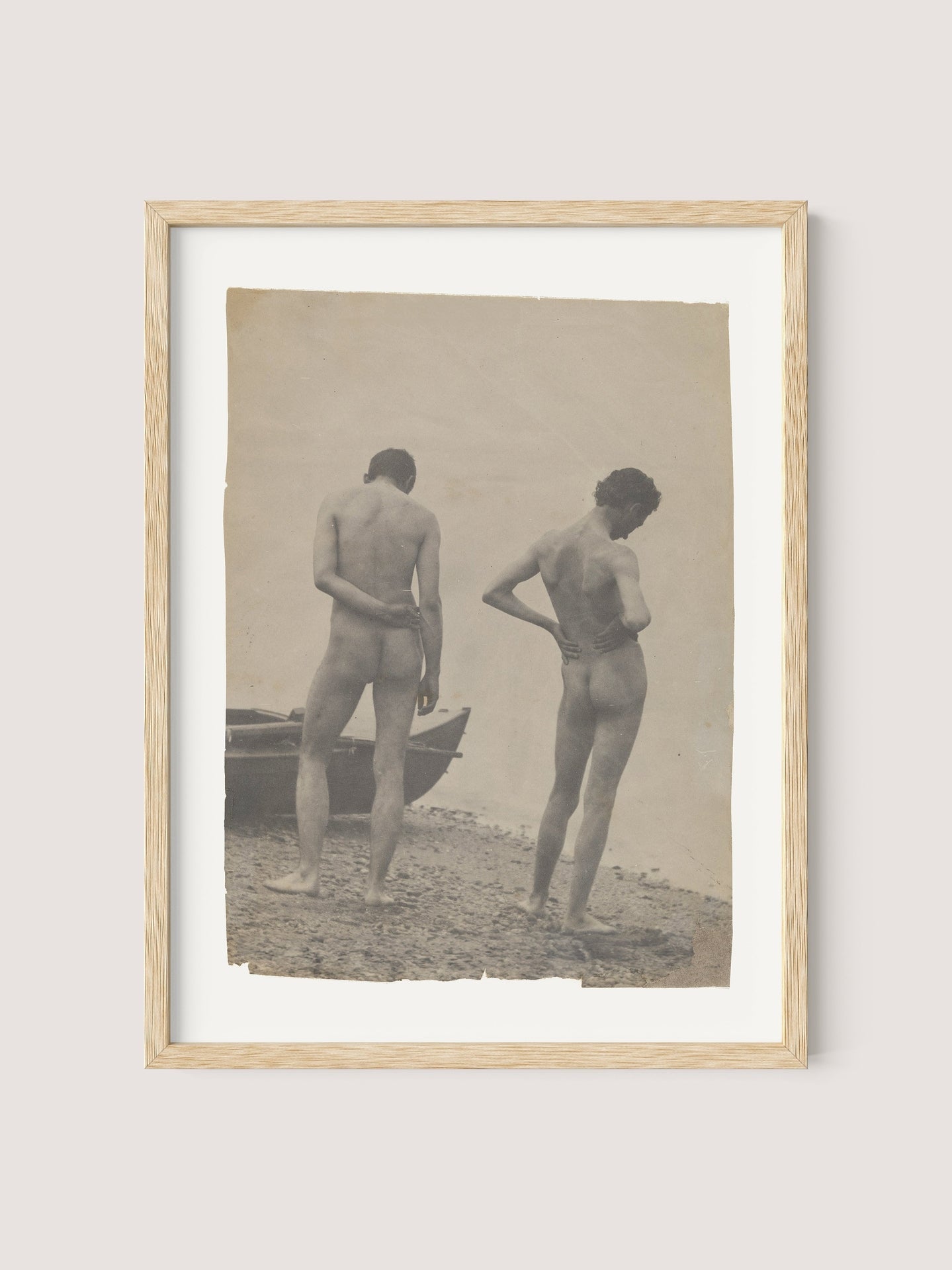 A framed photograph titled "Beach Walk," encased in natural oak, captures two nude figures standing on a rocky shoreline, reminiscent of Thomas Eakins' creative process. The figures, with their backs to the camera, appear contemplative; one stands upright while the other leans slightly with a hand resting on their lower back. A boat is visible to the left.