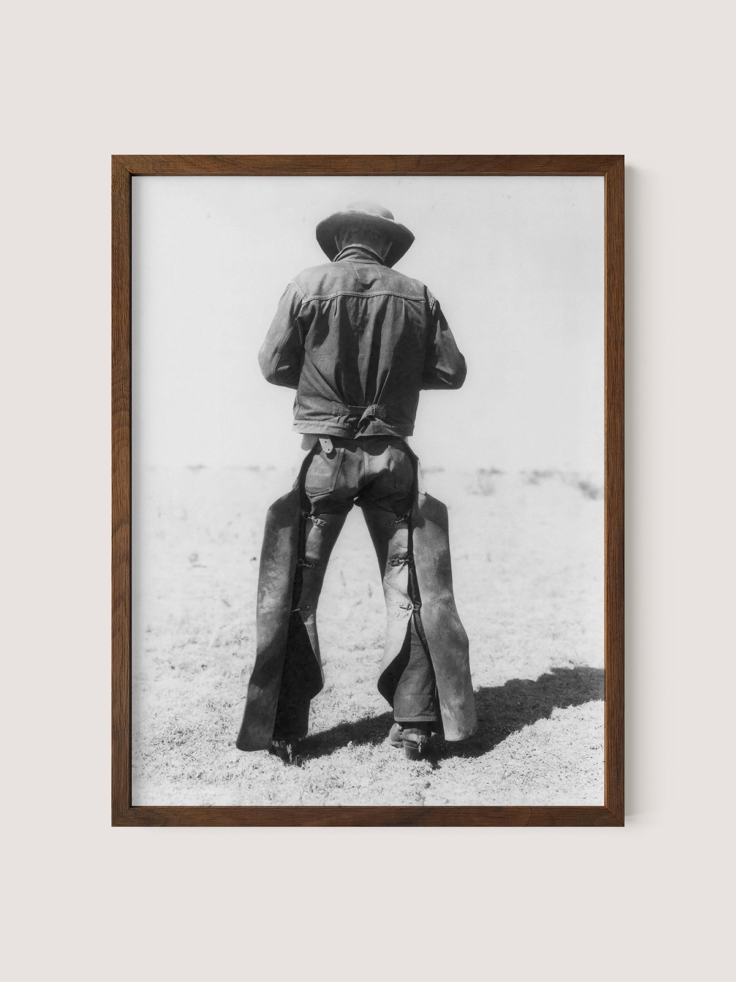 The &quot;Working Cowboy&quot; is a black and white print in a walnut oak frame, showcasing the back view of an American cowboy standing in a desolate landscape. The cowboy, adorned with a hat, jacket, and chaps, embodies a rugged and timeless western aesthetic that evokes a sense of nostalgia.