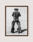 The "Working Cowboy" is a black and white print in a walnut oak frame, showcasing the back view of an American cowboy standing in a desolate landscape. The cowboy, adorned with a hat, jacket, and chaps, embodies a rugged and timeless western aesthetic that evokes a sense of nostalgia.